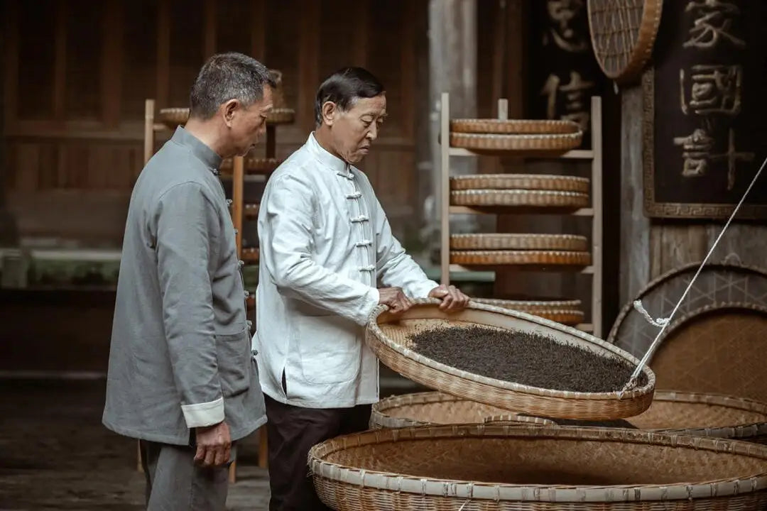 Two men examining tea leaves in a woven tray with Yixing teapots and Zisha clay craftsmanship