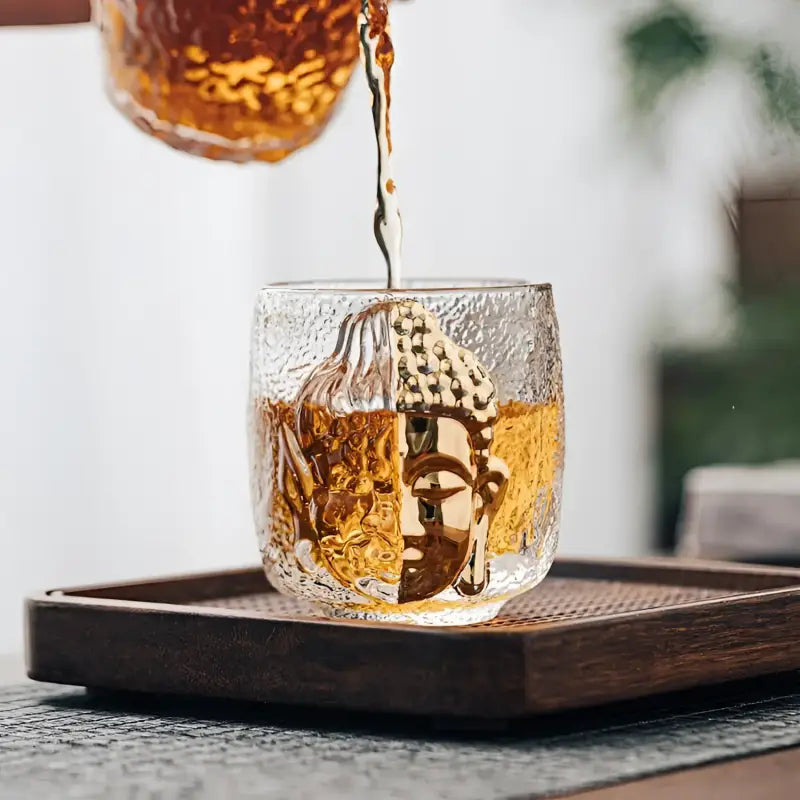 Buddha-faced glass cup with crackle glass teacup design being filled with tea, spiritual symbolism